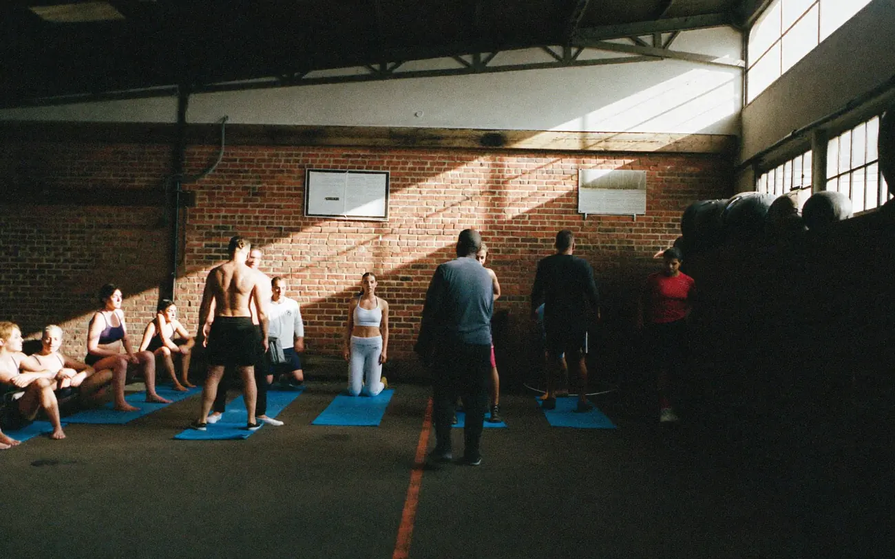 A busy gym floor with people warming up in rows on mats under high windows