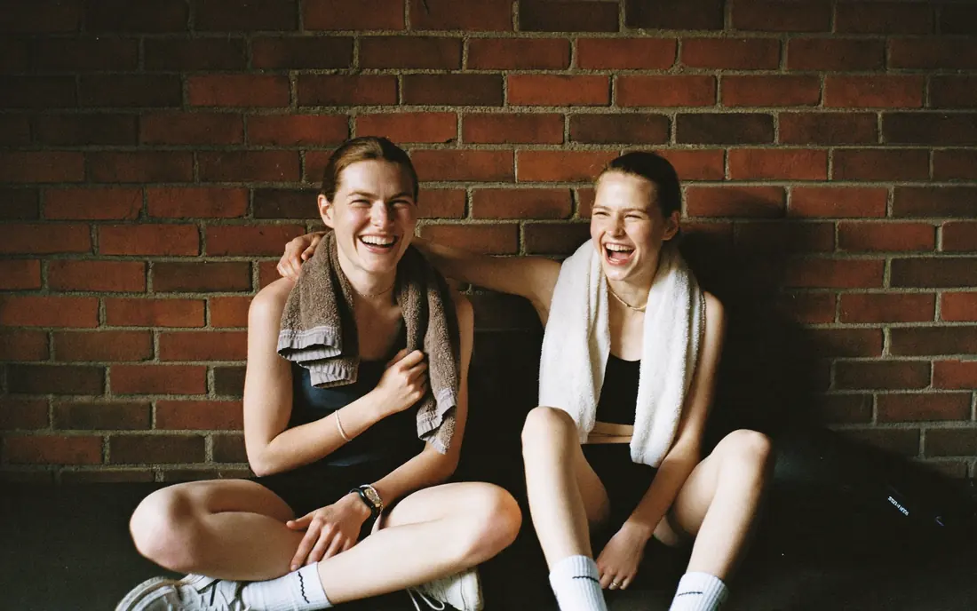 Two young women laughing against a brick wall after training