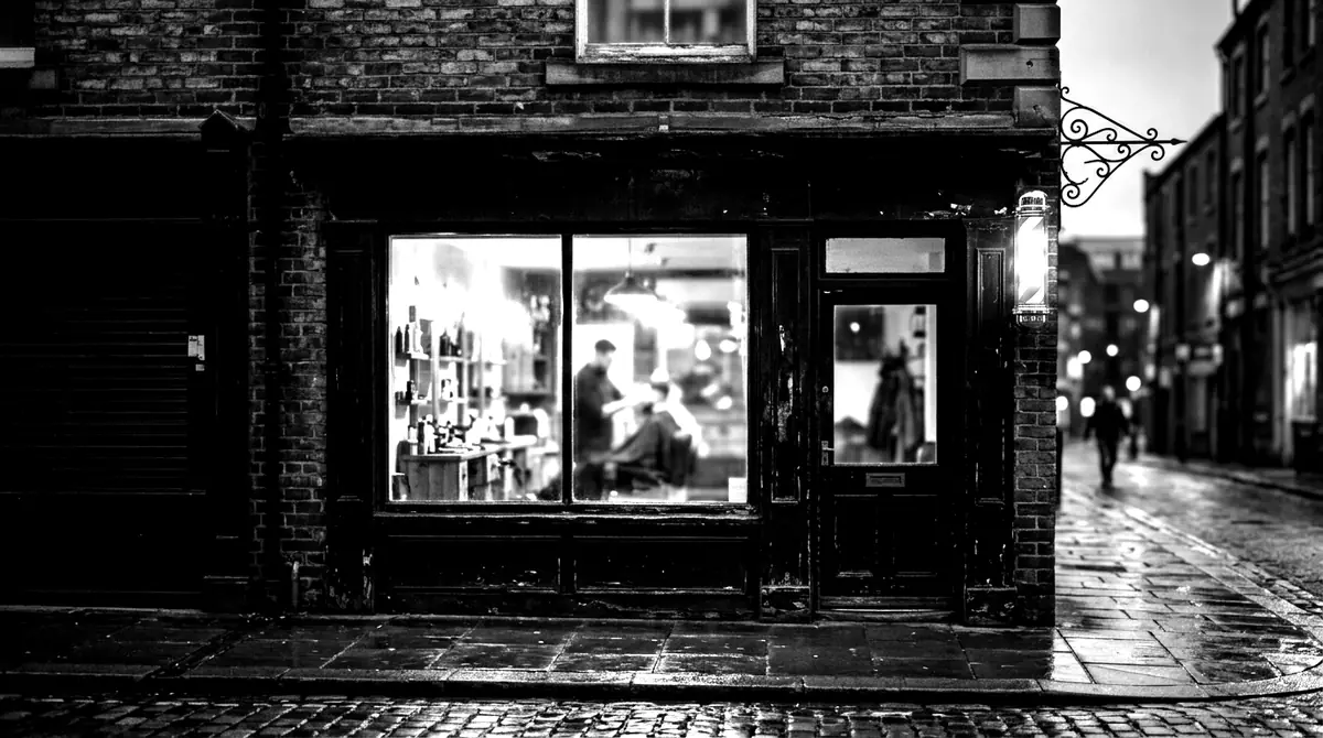 The near-black shopfront of Sharp House at dusk, window glowing warm, striped barber pole beside the door.
